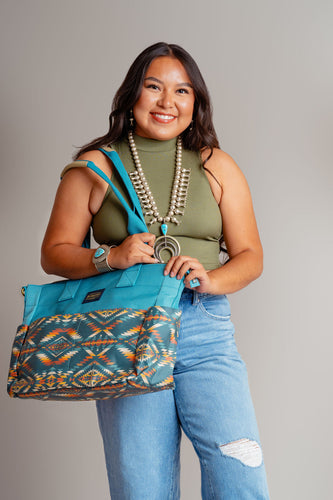 Young Navajo woman wearing blue jeans and a OD green top smiling and carrying a Pendleton blue tote bag on her right shoulder. She is standing in front of a beige background and wearing a squash blossom and large turquoise bracelet